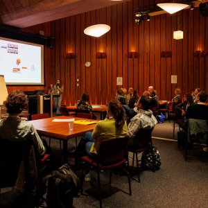 Around 20 participants are listening to a presentation. They are seated around a number of square tables, and the presentation is projected onto a screen.