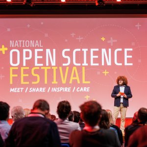 The chairperson leads the festival’s closing session, standing on the stage in the auditorium. In the background, the logo is displayed in prominent view on the projection screen.